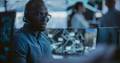 African Specialist in a Dark High-Tech Monitoring Room, Concentrated on a Call and Computer Work....