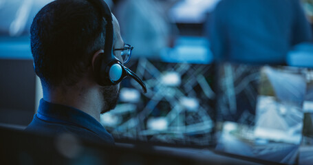 Technical Customer Support Specialist is Talking on a Headset while Working on a Computer in a Call Center Control Room Filled Display Screens and Data Servers. Footage from the Back