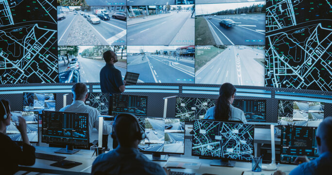 Male Software Engineer Working on Laptop Next to a Big Screen in a Modern Monitoring Office with Live CCTV Footage with Traffic Situation. Monitoring Room With Big Data Engineers Work on Computers