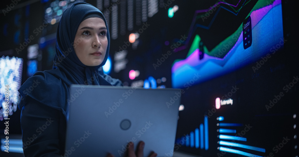 Poster Muslim Female Data Center IT Engineer Standing in a Room with Statistics and Charts Settings on a Digital Screen. Cloud Computing Expert Uses Laptop for Servicing the System in Cyber Security Facility
