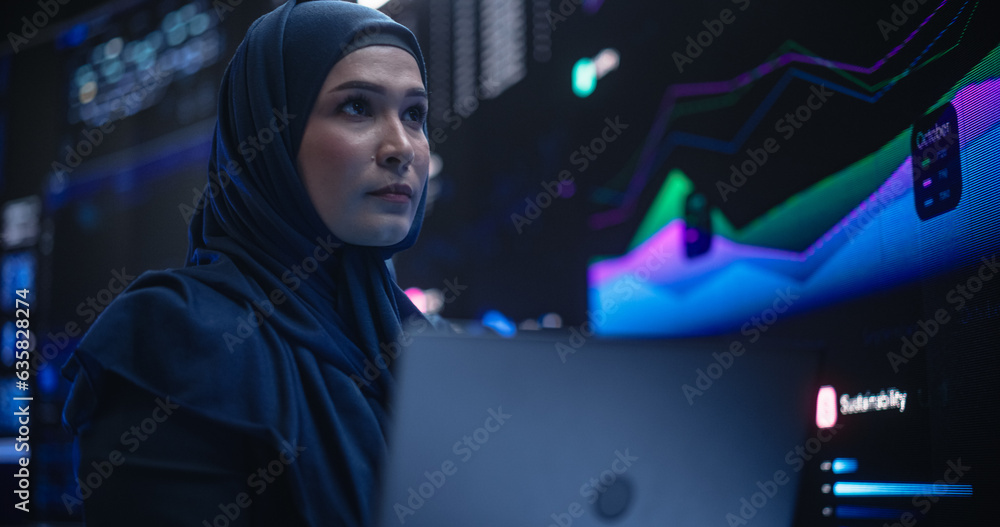 Poster Muslim Female Data Center IT Engineer Standing in a Room with Statistics and Charts Settings on a Digital Screen. Cloud Computing Expert Uses Laptop for Servicing the System in Cyber Security Facility