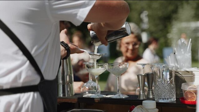 Bartender at party with people pours cocktail from shaker through strainer into wide glass. Work of professional on sunny day and straining drink through sieve on bar counter outdoor.