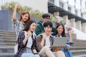 Group of Asian students with raised arms celebrate celebrating completion of exams through laptop computer.