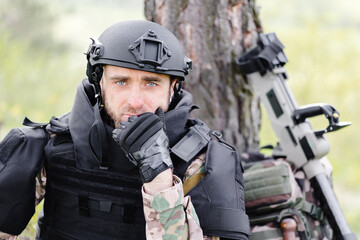 A man in a military uniform and a bulletproof vest sits in the forest near a metal detector and a military backpack. A man pauses in the work of demining the territory