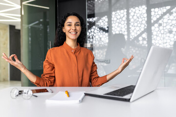 Young successful businesswoman meditating at workplace, Hispanic woman smiling and looking at camera in lotus position at workplace using laptop.