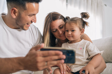 Father and mother showing drawings on the mobile phone to entertain their young daughter sitting on...