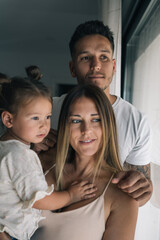 Mother holding her young daughter in her arms while her father grabs the woman by the shoulder and they all look out the window.