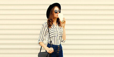 Portrait of stylish young woman drinking coffee looking away wearing black round hat, sunglasses, striped shirt on white background