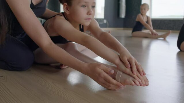 Medium shot of little gymnast trying to do difficult stretching exercise with legs while sitting with teacher on floor
