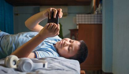 Happy Asian kid boy playing with smartphone lying on a cozy bed in a room at home