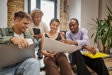 happy interracial team holding gadgets, sitting on stairs in coworking, looking at startup project