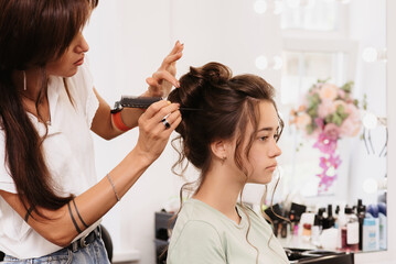 Shooting in a beauty salon. The hair master corrects the hairstyle of a young dark-haired girl with the help of a hair brush.