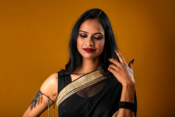 Portrait of a young beautiful girl wearing traditional black saree posing on a brown background.