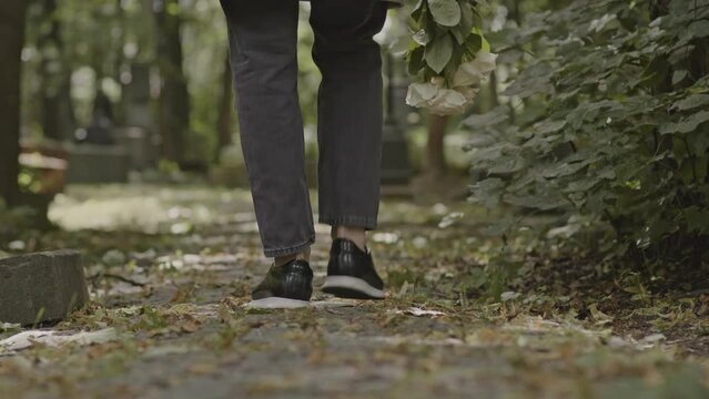 Back view slowmo of widower with bouquet of white roses walking on cobblestone graveyard alley to grave of his beloved wife