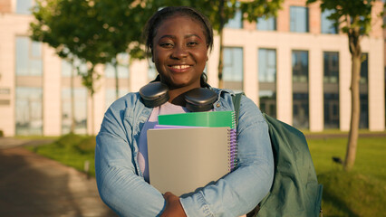 Fototapeta premium Portrait African American woman university college campus academy student teen girl high school pupil holding books with schoolbag headphones smiling toothy joyful in city outdoors study education