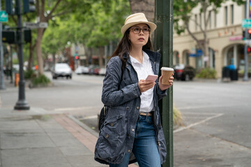 asian Japanese female visitor wearing hat and carrying coffee using navigation app on phone on the street while touring palo alto city in California usa in spring