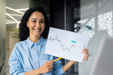A businesswoman at an online meeting looks into the laptop camera and shows her colleagues and partners a graph of financial income, a satisfied financier at a startup presentation inside the office