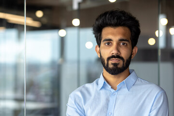 Close-up photo. Portrait of a young Indian businessman man wearing a blue shirt standing in the office and seriously looking at the camera