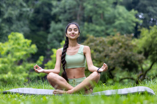 Beautiful young Indian woman doing yoga in the park. Sitting on a mat with closed eyes in the lotus position and meditating
