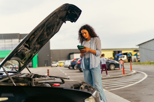 A young woman stands in front of a car with the hood up and reads the instructions on how to change the oil