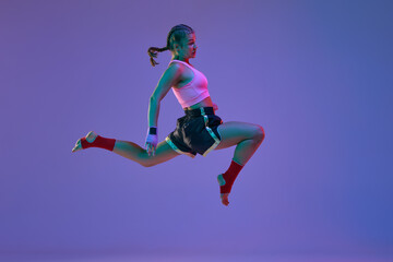 Kick in a jump. Teen girl, mma athlete in motion, training against purple studio background in neon lights. Concept of mixed martial arts, sport, hobby, competition, athleticism, strength, ad