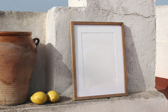 Blank vertical wooden frame picture mock up with vintage olive clay storage pot, vase. White old textured white wall in sunlight. Lateral view, no people. Summer display background for art, posters.