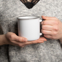 Closeup of female hands with a ceramic mug of beverage. Beautiful girl holding a white black cup of tea or coffee in the morning sunlight. Mug for your design. Empty mockup template 