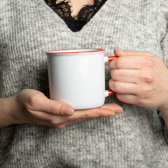 Closeup of female hands with a ceramic mug of beverage. Beautiful girl holding white red cup of tea or coffee in the morning sunlight. Mug for your design. Empty mockup template 