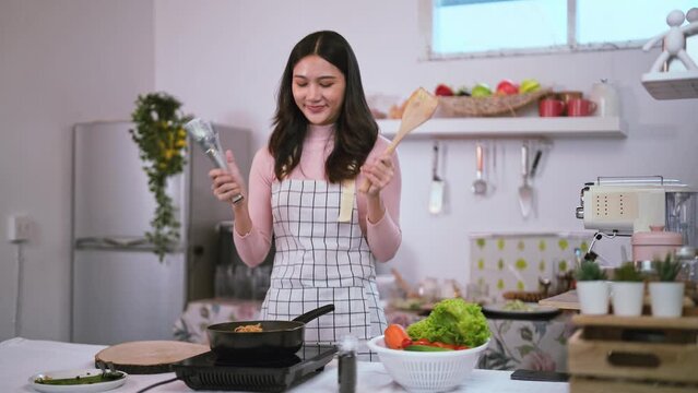 Asian young woman wearing apron dancing looks happy while cooking in the kitchen at home, happiness expression in lifestyle