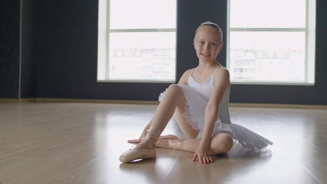 Portrait of positive small ballerina sitting on floor of dance studio in ballet-clothes and looking at camera in panoramic window background