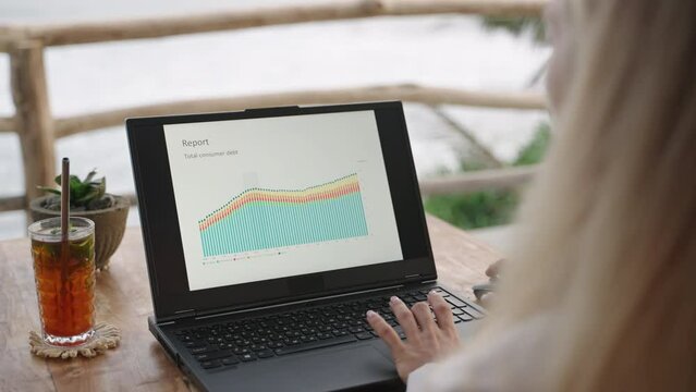 Woman watching presentation slides on laptop screen sitting in tropical cafe with sea view and palm trees. Female designer making presentation on computer at coworking. Remote work, business.