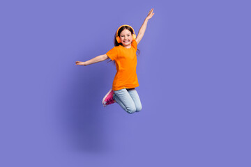Fototapeta na wymiar Full length photo of cheerful schoolkid dressed orange t-shirt flying in headphones arms wings isolated on purple color background