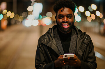 Portrait of a happy urban African American man standing on the city street at night and typing on...