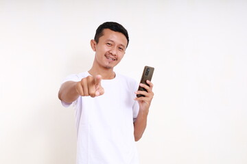 Happy asian funny man holding a cell phone while pointing at you. Isolated on white background