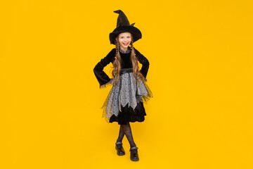 A child in a witch costume. Carnival for children on autumn Halloween. A little girl in a pointed hat and dress is smiling broadly. Happy holiday. Bright yellow isolated background.