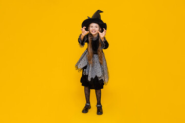 A child in a witch costume. Carnival for children on autumn Halloween. A little girl in a pointed hat shows portrays an evil witch. Happy holiday. Bright yellow isolated background.