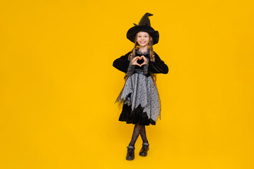 Happy smiling baby on Halloween. A full-length schoolgirl in a carnival witch costume. A young girl shows a heart with her fingers. A little fairy girl on a bright orange isolated background.