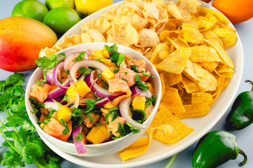 Salmon Ceviche with Mango Served with Plantain and Tortilla Chips Close-up: A bowl of salmon and mango ceviche with chips surrounded with ingredients