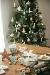 Stylish christmas gift with white ribbon and modern decorations on wooden table on background of festive decorated tree in scandinavian room. Merry Christmas and Happy holidays!