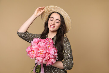 Beautiful young woman in straw hat with bouquet of pink peonies against light brown background