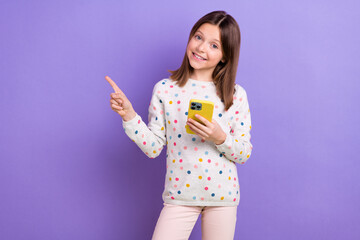 Portrait of charming cheerful schoolkid hold smart phone direct finger empty space promo isolated on purple color background