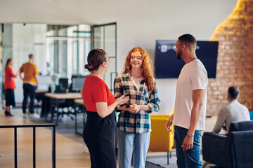 A group of young business individuals, including a girl with orange hair and an African American man, stands in a modern corporate hallway, collectively examining business progress on a smartphone