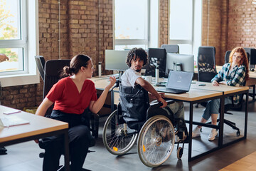 A young business group, including an African American businessman in a wheelchair, collaborates within a modern glass office, actively engaged around a computer and laptop, collectively solving