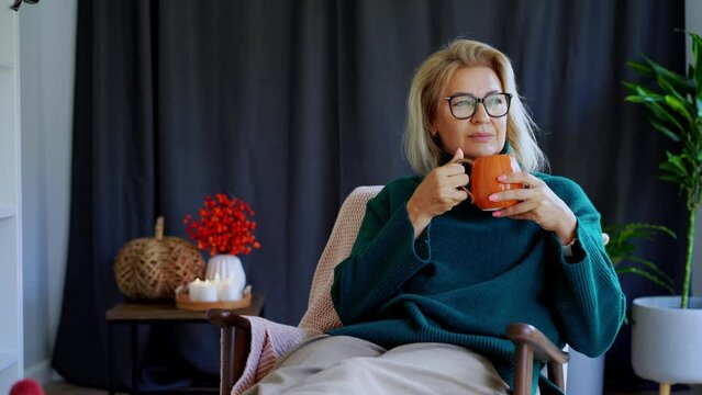 Middle aged woman relaxing with pumpkin shaped cup of hot drink in scandy style hygge interior home with fall mood decor. Lady dreaming, enjoy calm mood without stress, well being alone. Cozy autumn