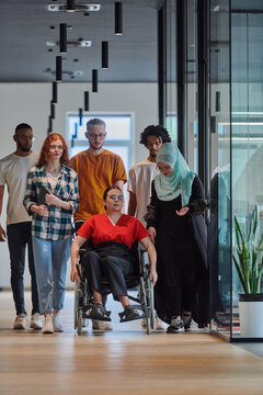 A diverse group of young business people walking a corridor in the glass-enclosed office of a modern startup, including a person in a wheelchair and a woman wearing a hijab, showing a dynamic mix of