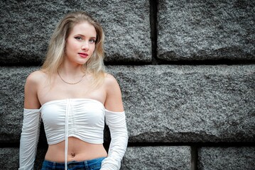 Blonde girl wearing a white top and blue jeans, standing against a stone wall.