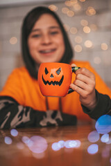 Portrait of candid authentic zoomer boy posing with jack lantern mug and sweets at halloween party