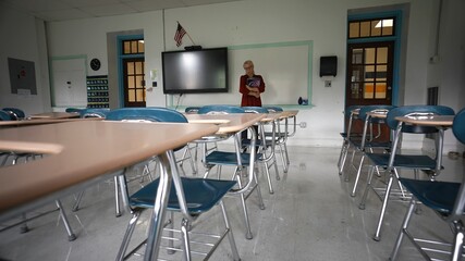 Sad unhappy teacher in large empty classroom with the lights turned off during budget cuts or a pandemic wondering about the future of education.