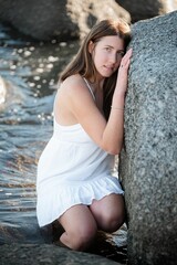 Brunette girl in a white sundress crouching in a pool of water and leaning against a boulder