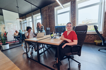 A young business group, including an African American businessman in a wheelchair, collaborates within a modern glass office, actively engaged around a computer and laptop, collectively solving